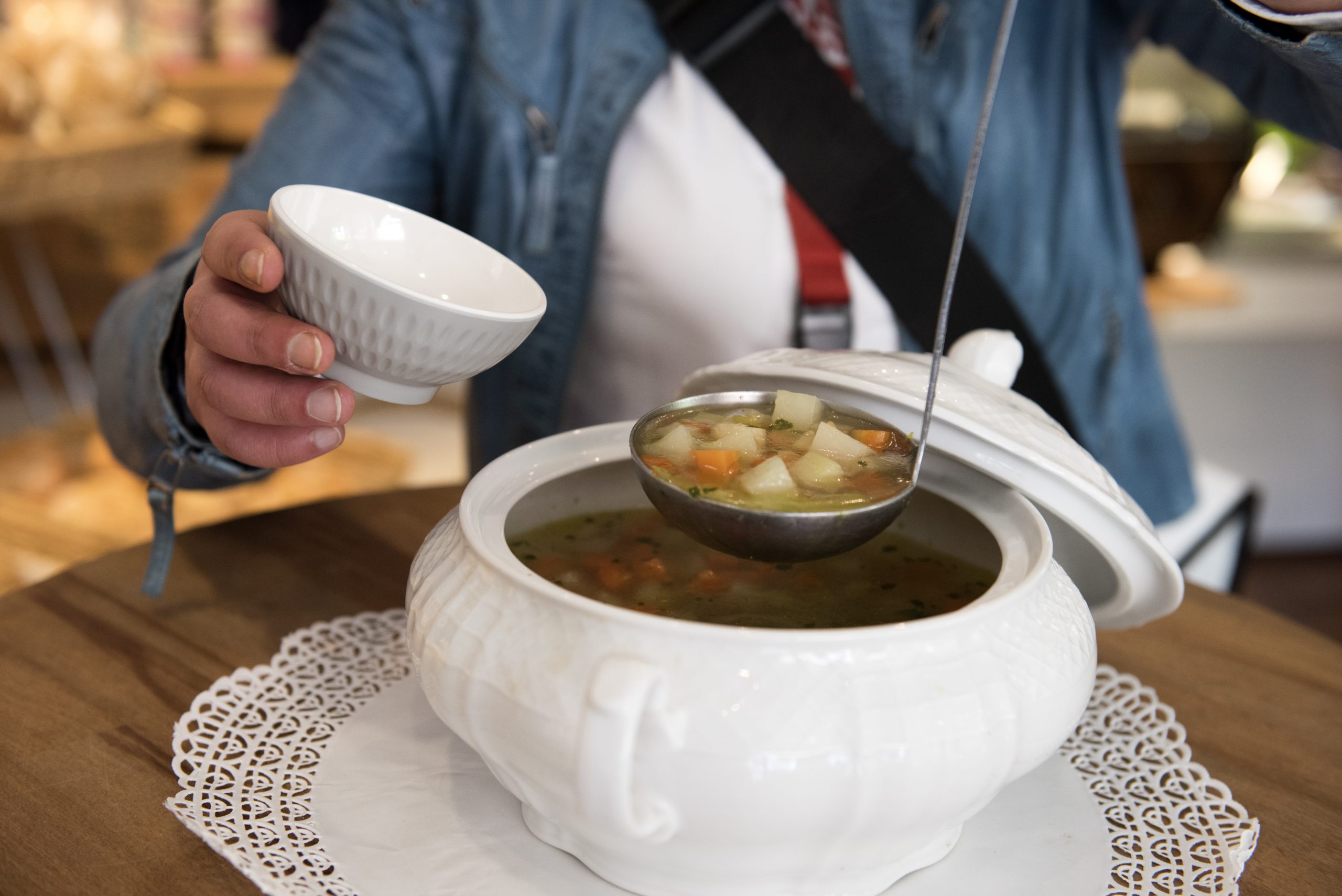 Eine Person serviert elegant Gemüsesuppe aus einer weißen Terrine in eine Schüssel auf einem Holztisch und fängt damit den Geist der Erlebnisgastronomie ein.