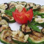 Gegrillte Zucchinischeiben mit Knoblauch und Petersilie, garniert mit einer Tomatenblüte auf einem weißen Teller, bieten ein Erlebnisgastronomie-Erlebnis, das die Sinne verwöhnt.