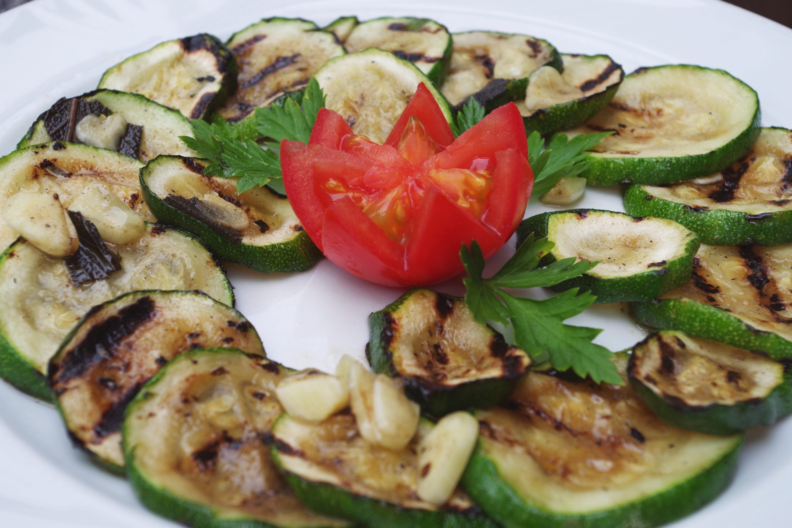 Gegrillte Zucchinischeiben mit Knoblauch und Petersilie, garniert mit einer Tomatenblüte auf einem weißen Teller, bieten ein Erlebnisgastronomie-Erlebnis, das die Sinne verwöhnt.