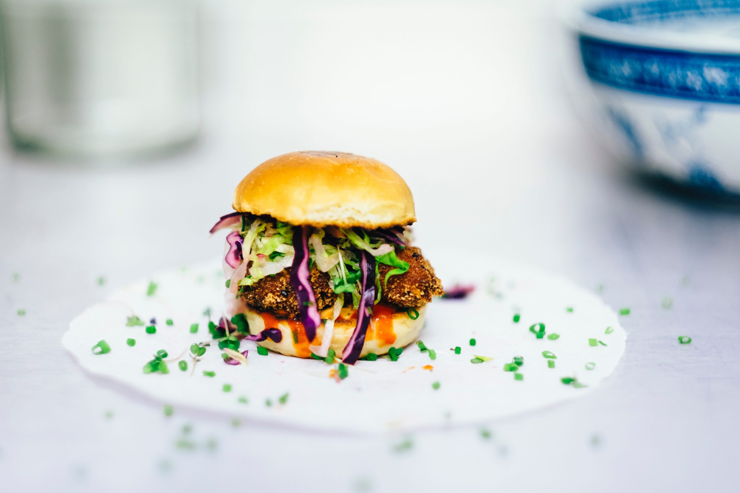 Mini-Slider-Burger mit frittiertem Patty, buntem Krautsalat und Kräutern, serviert als genussvolles Beispiel der Erlebnisgastronomie auf einem weißen Teller, mit einer unscharfen Schüssel im Hintergrund.