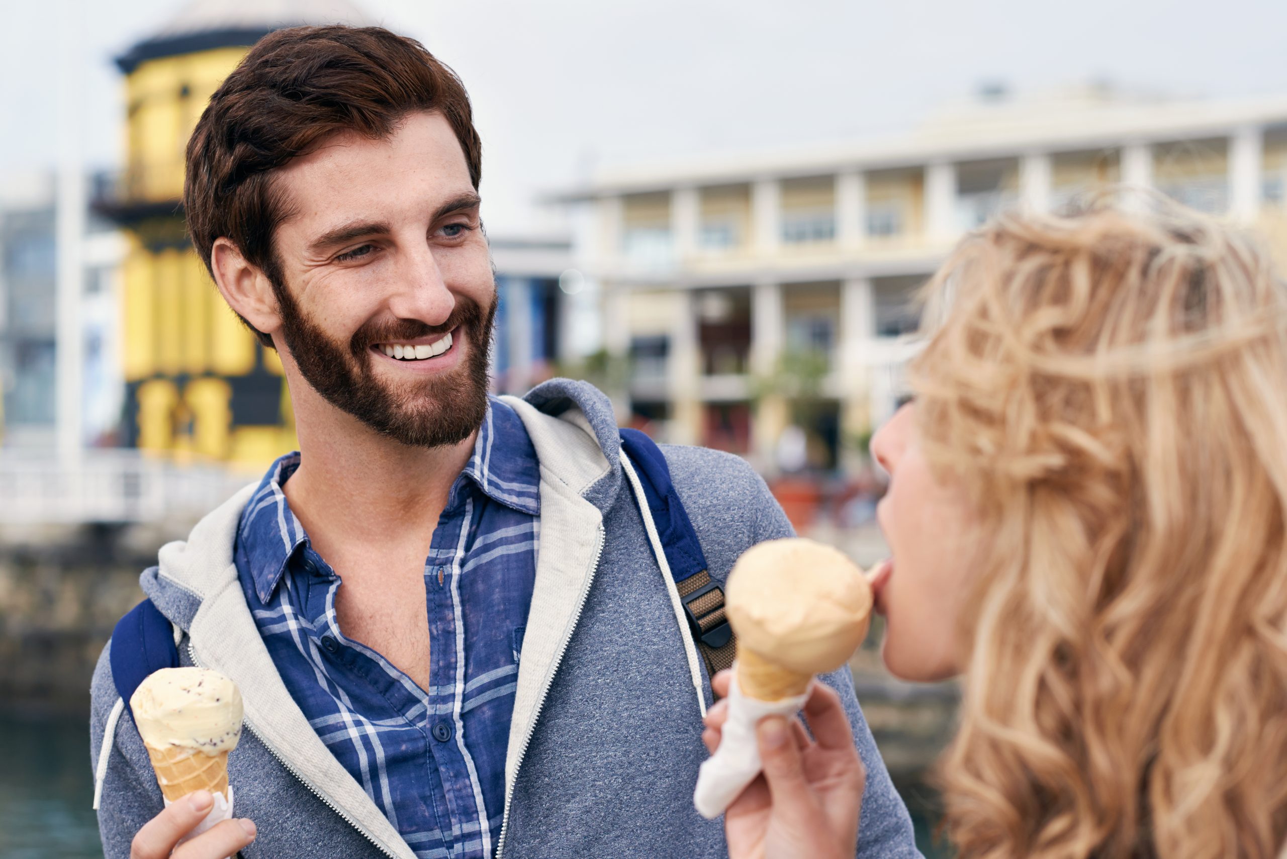 Ein lächelnder Mann mit Bart und eine Frau genießen Erlebnisgastronomie mit Eiswaffeln in der Nähe einer Uferpromenade, im Hintergrund ist ein gelber Turm zu sehen.