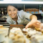 Eine lächelnde Frau genießt die Erlebnisgastronomie, während sie in der Vitrine einer Bäckerei nach verlockendem Gebäck greift.