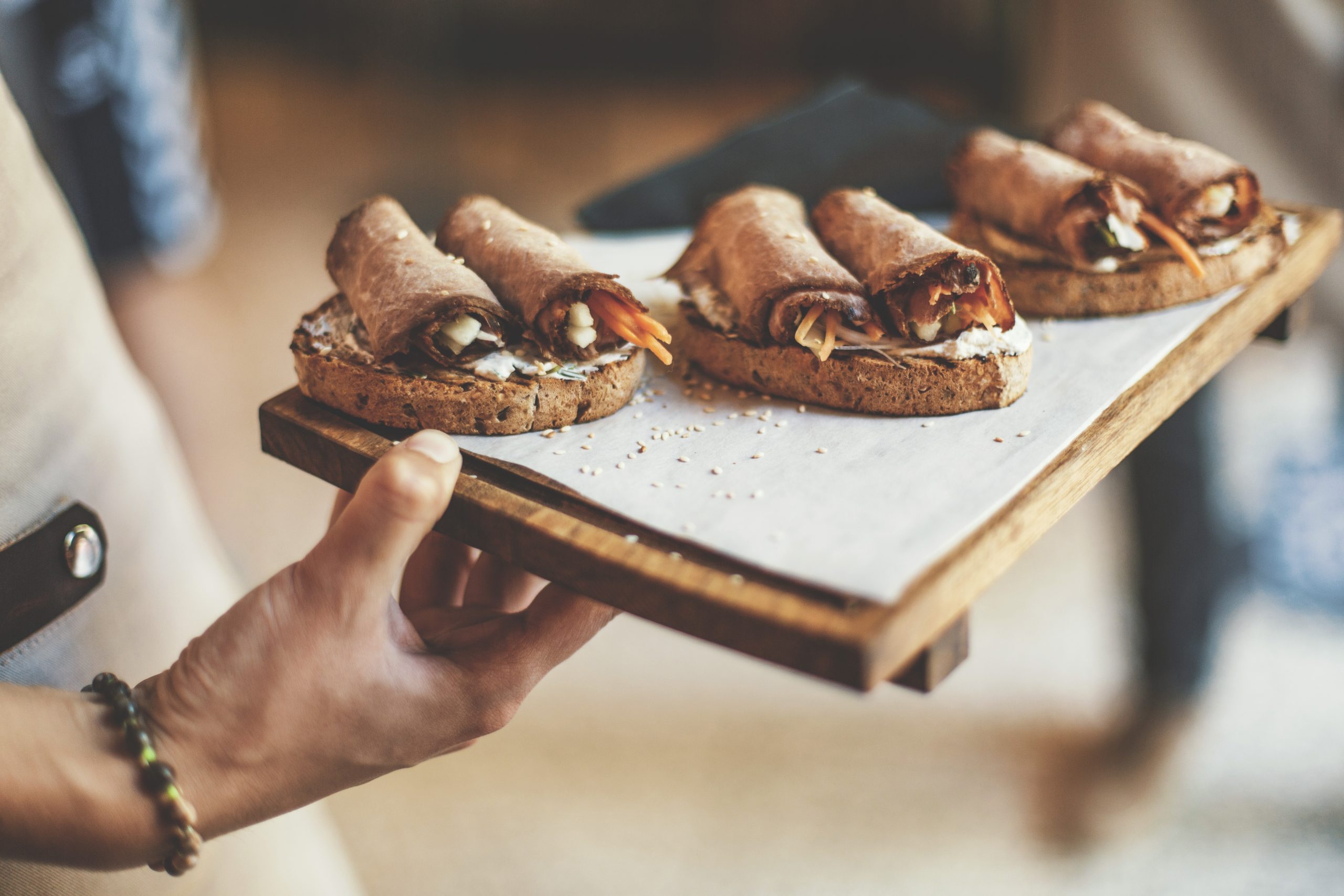 Eine Person hält ein Tablett mit Brot, das mit gerolltem Wurstaufschnitt und Gemüse belegt ist, und bietet ein herrliches Erlebnisgastronomie an.