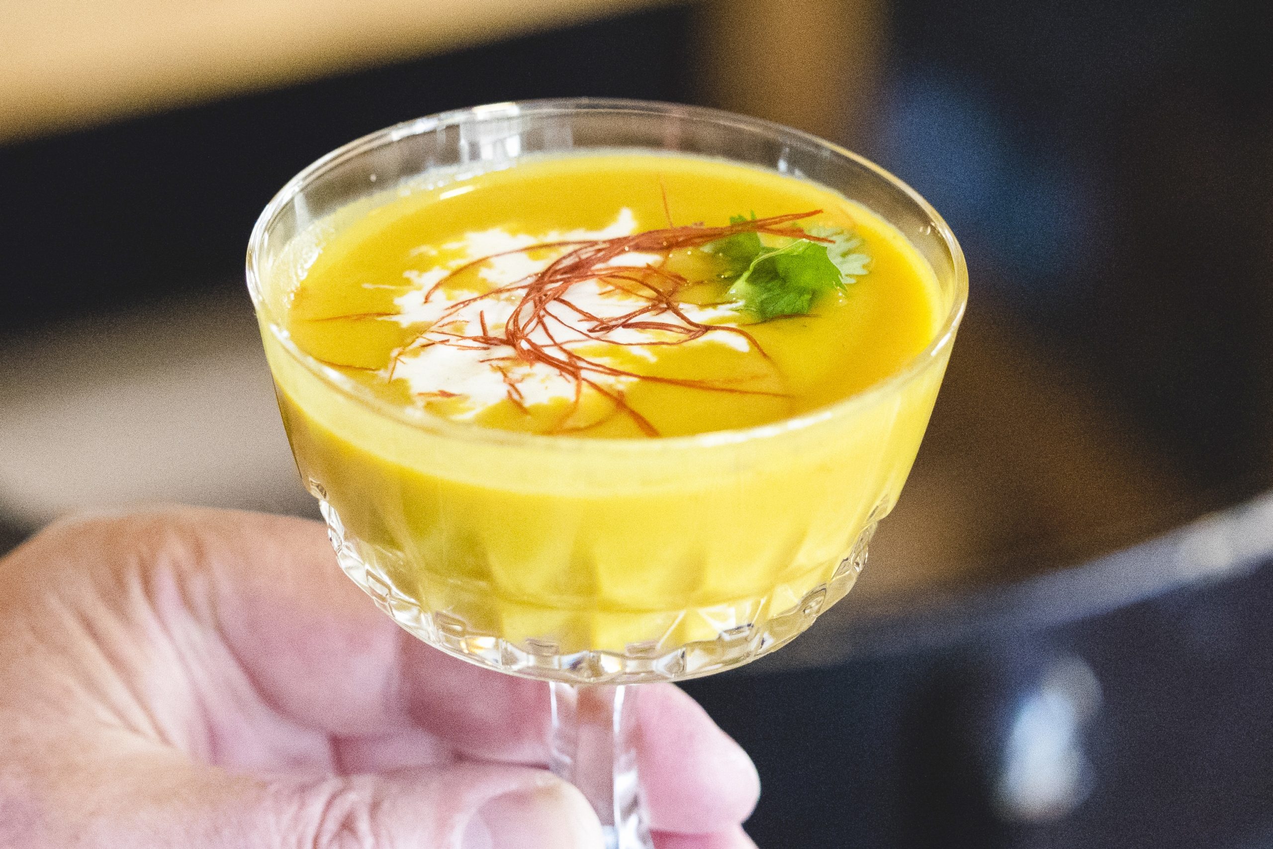 Eine Hand hält ein Glas cremige Orangensuppe, garniert mit Sahne, Kräutern und feinen roten Fäden – ein exquisiter Geschmack der Erlebnisgastronomie.