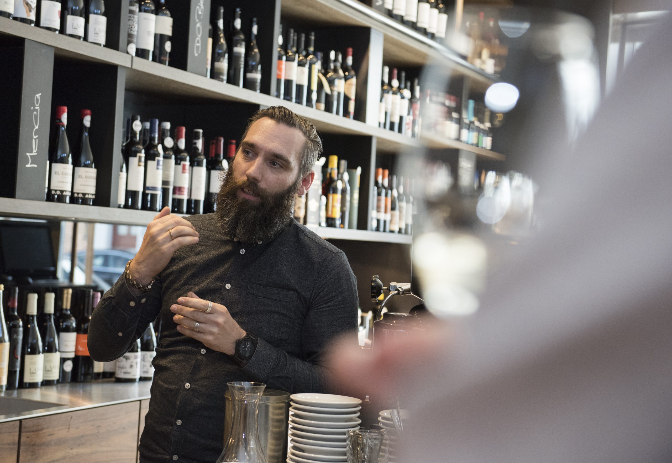 Ein bärtiger Mann gestikuliert leidenschaftlich, während er in einem Weinladen über die Auswahl erlesener Weine spricht und inmitten von Regalen voller erlesener Flaschen die Essenz der Erlebnisgastronomie verkörpert.