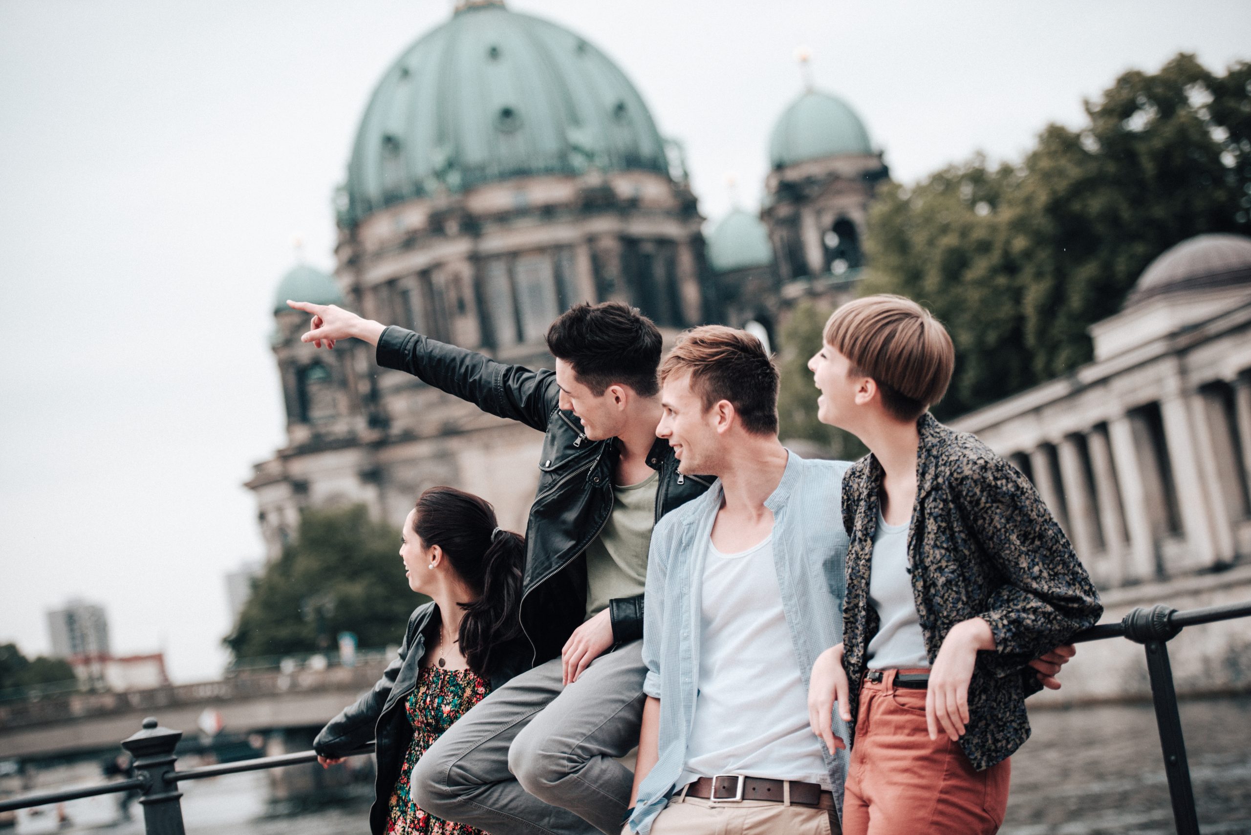 Vier Menschen sitzen lächelnd auf einem Geländer und genießen ein Erlebnis, das Essen und Unterhaltung vereint, mit einem Kuppelgebäude im Hintergrund.