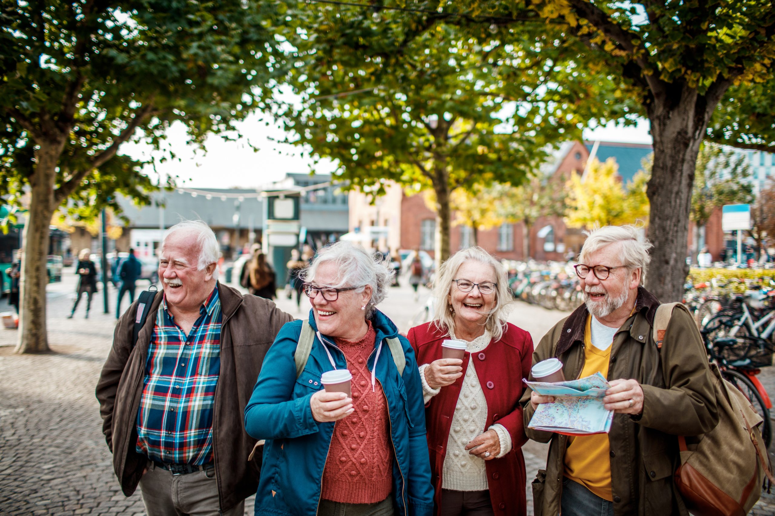 Vier ältere Freunde lachen mit Kaffee und Karte in einer lebhaften städtischen Umgebung und entdecken bei einem Spaziergang unter den Bäumen neue Erfahrungen, die einem erlebnisgastronomischen Abenteuer ähneln.