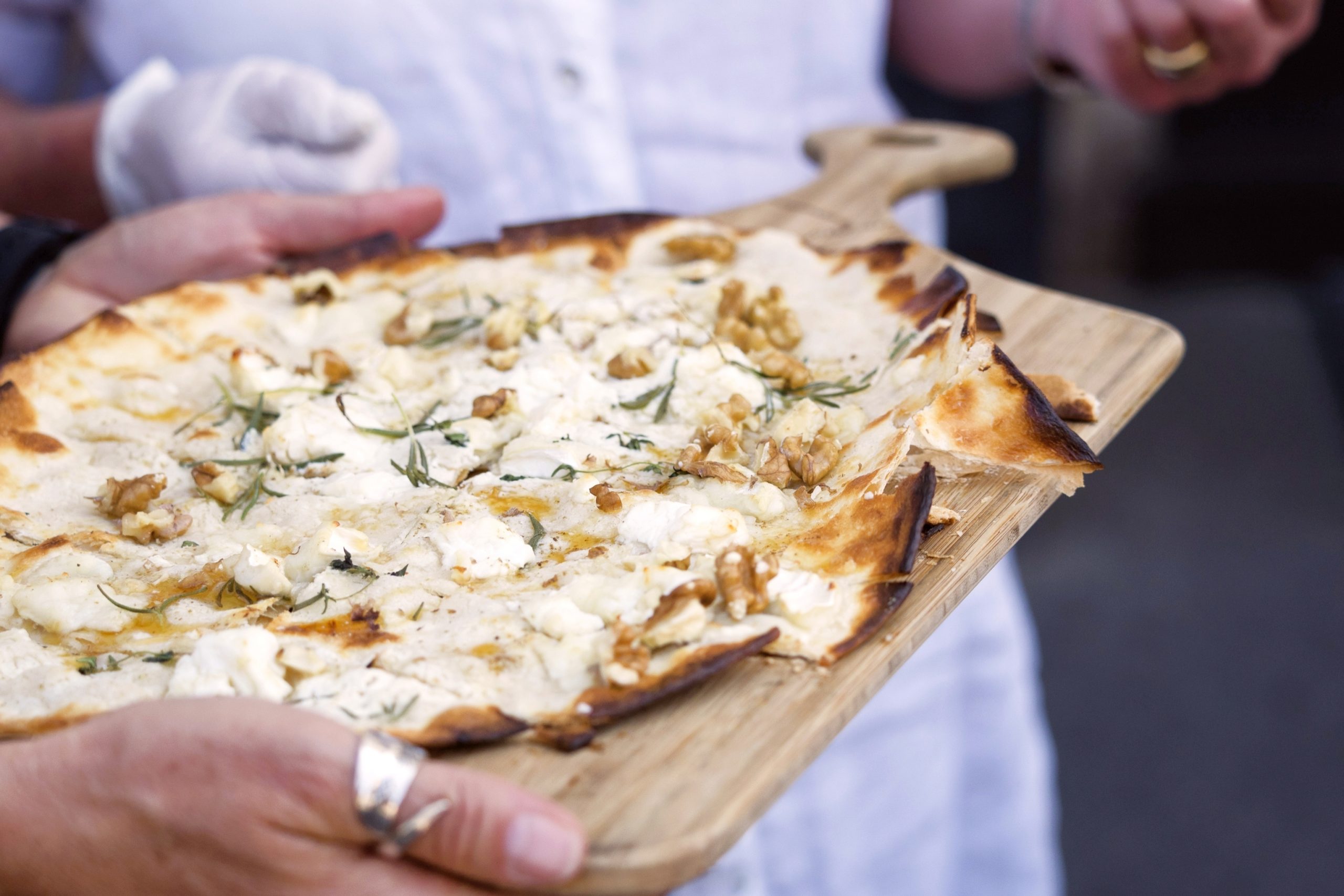 Eine Person hält ein Holzbrett, auf dem Erlebnisgastronomie vom Feinsten präsentiert wird: rustikales Fladenbrot mit Käse, Kräutern und Walnüssen belegt.