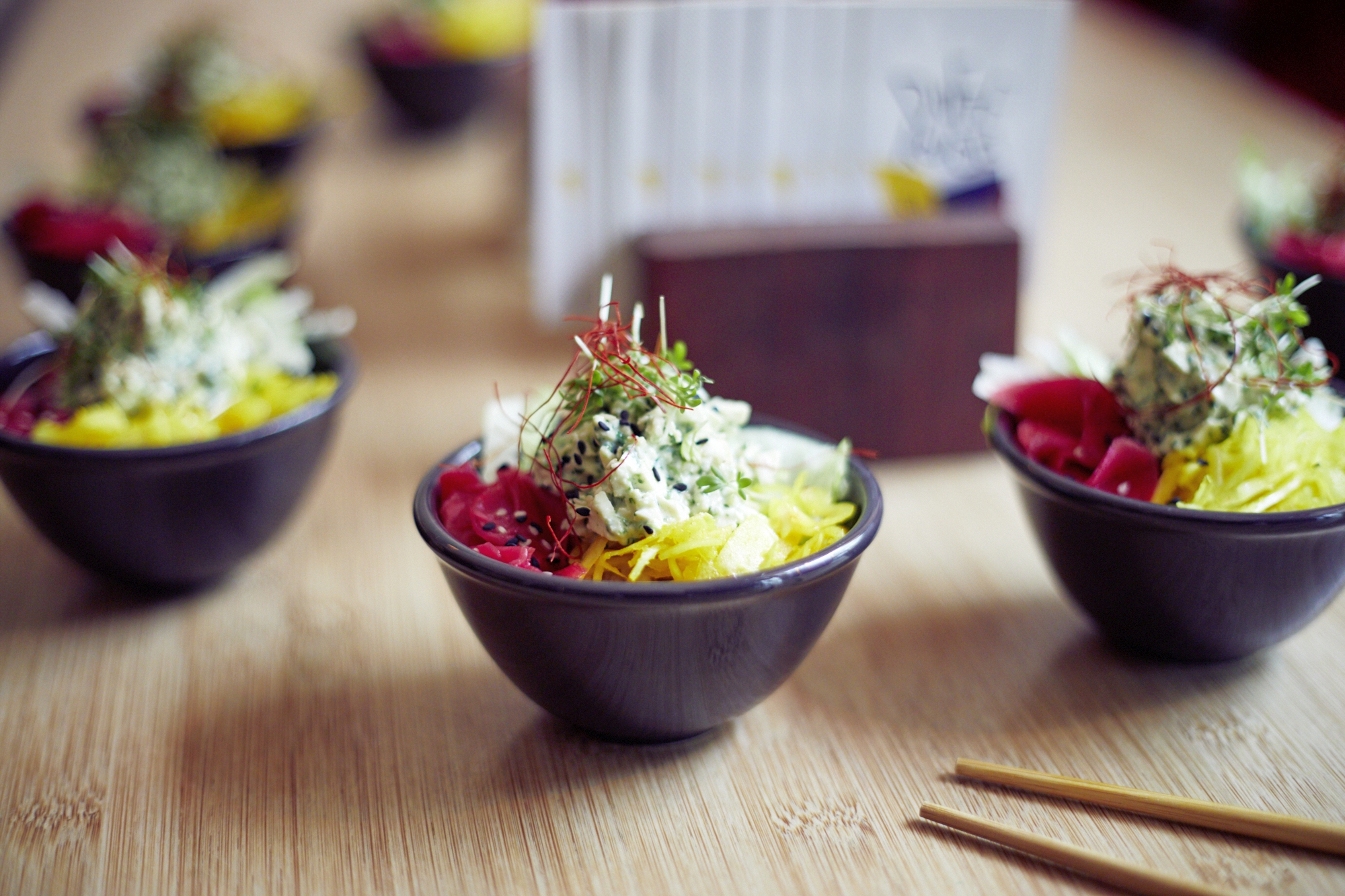 Kleine Schüsseln mit Salat, Sprossen und geraspeltem Gemüse stehen auf einem Holztisch, daneben sind kunstvoll Essstäbchen platziert und bieten ein erlebnisgastronomisches Erlebnis, das die Sinne erfreut.