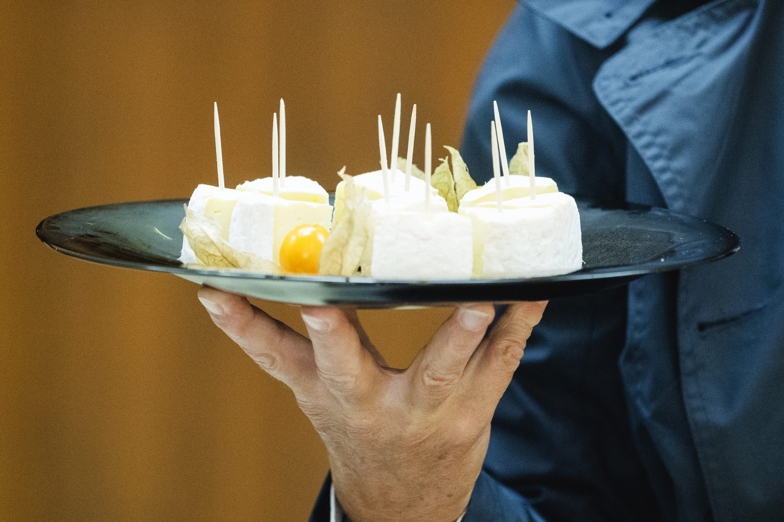 Eine Person präsentiert einen Teller mit Scheibenkäse, Zahnstochern und einer kleinen gelben Tomate und lädt Sie zu einem erlebnisgastronomischen Erlebnis ein.