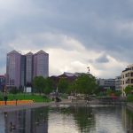 Stadtparkszene mit einem Teich, Gänsen, hohen Gebäuden und einem bewölkten Himmel.