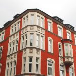 Ein historisches Gebäude in Rot und Beige mit großen Fenstern und kunstvollen Details unter einem bewölkten Himmel.