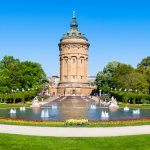 Panoramablick auf einen historischen Wasserturm, umgeben von Gärten und Springbrunnen unter einem klaren blauen Himmel.