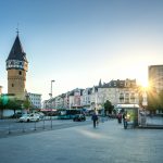 Stadtplatz bei Sonnenuntergang mit einem hohen Turm und Gebäuden, spazierenden Menschen und einem sichtbaren U-Bahn-Eingang.