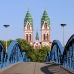 Eine blaue Eisenbrücke führt zu einer Kathedrale mit Zwillingstürmen mit grünen Dächern, vor einem klaren blauen Himmel.