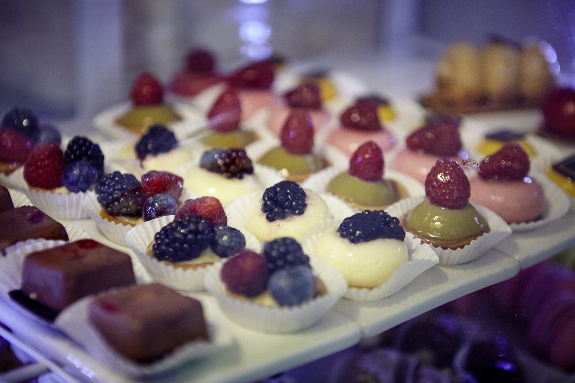 Verschiedene Mini-Desserts mit Beeren und buntem Topping, perfekt als köstliche Geschenkidee, präsentiert in einer Vitrine.