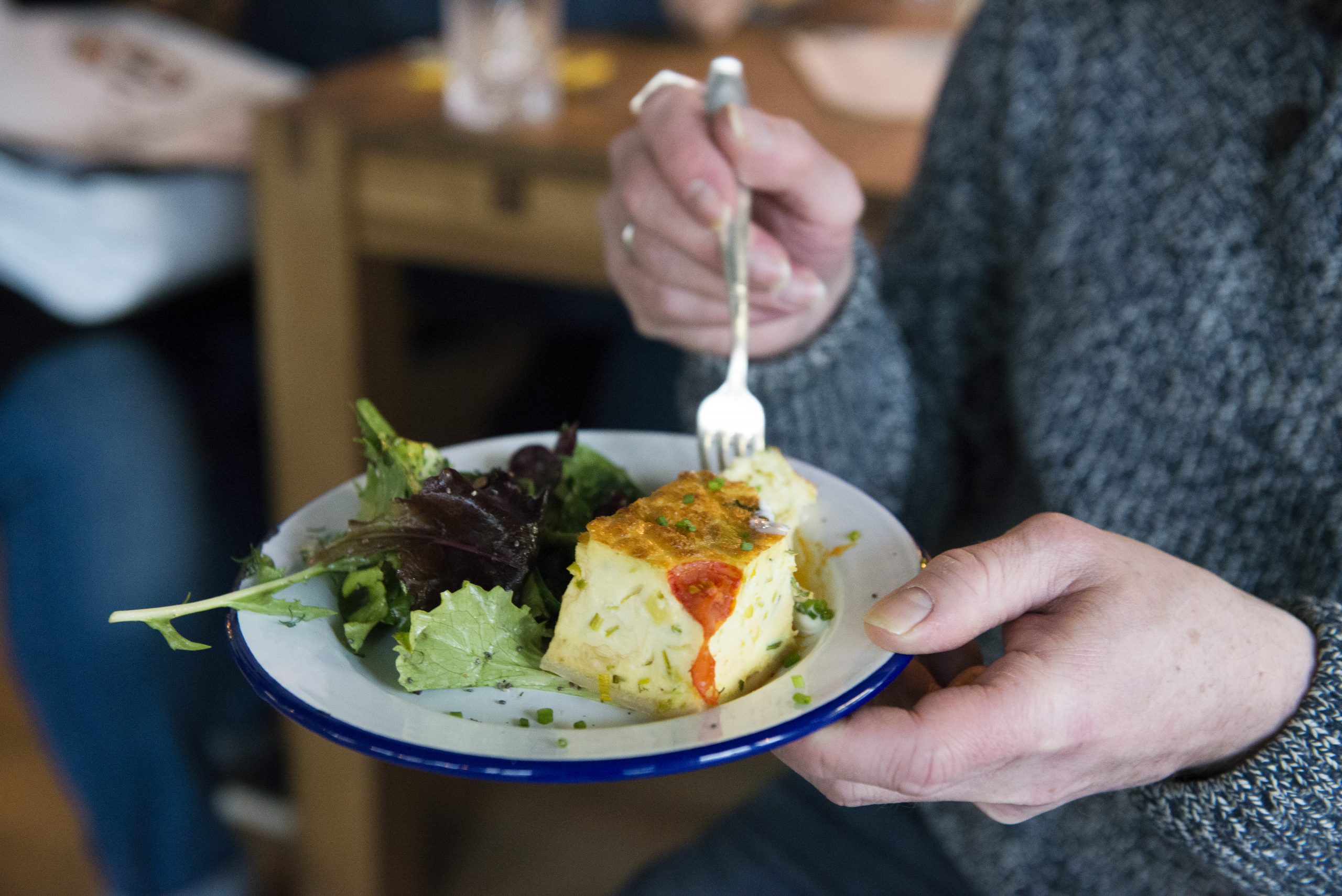 Eine Person hält einen Teller mit einem Stück Quiche und gemischtem Blattgemüse und nimmt mit einer Gabel einen Bissen – eine köstliche Mahlzeit, die auch die perfekte Geschenkidee für jeden Feinschmecker sein könnte.