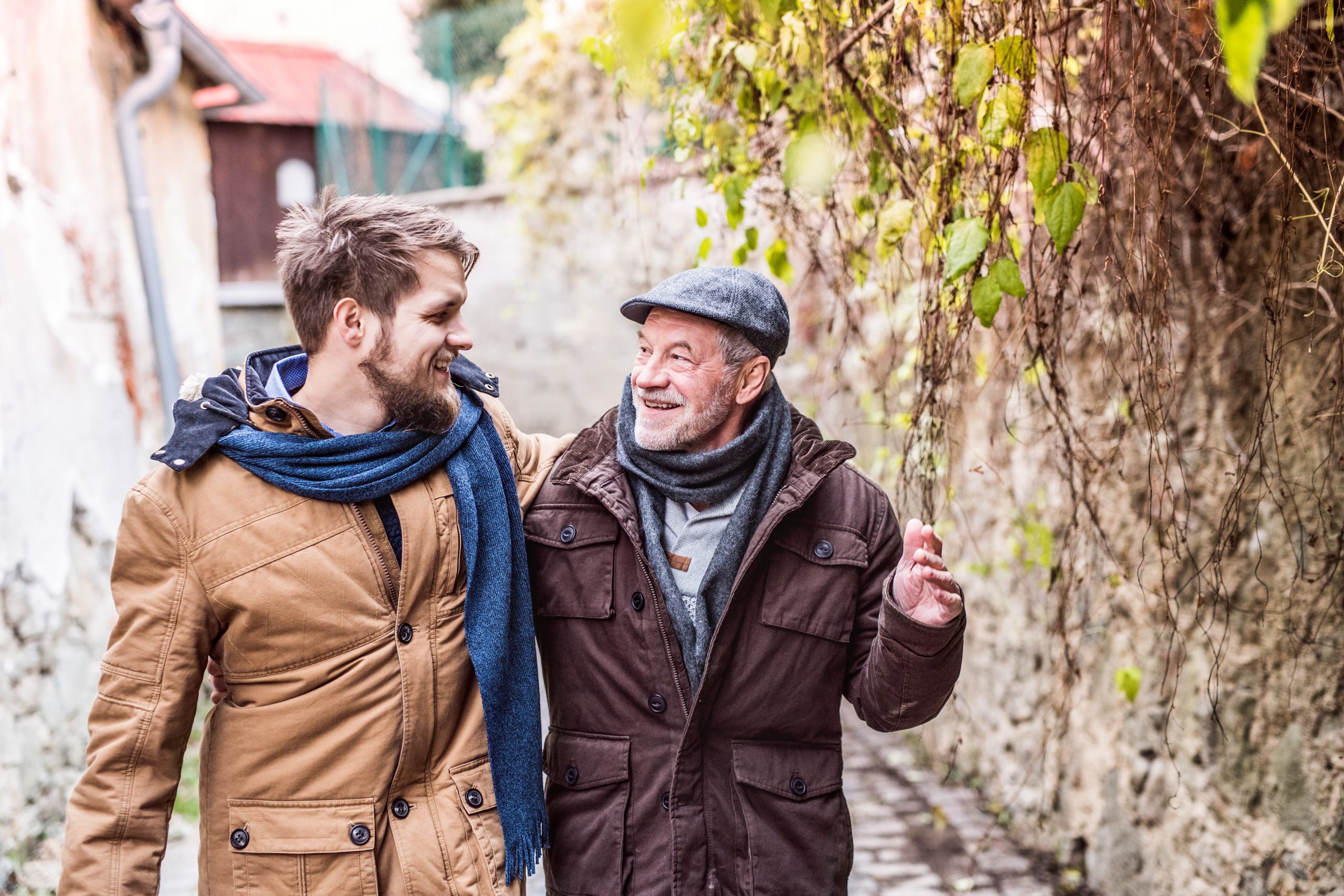 Zwei Männer, in Mäntel und Schals gehüllt, lächeln, während sie an einem Tag im Laub einen Kopfsteinpflasterweg entlangschlendern – vielleicht beim Brainstorming zu ihrer nächsten Geschenkidee.