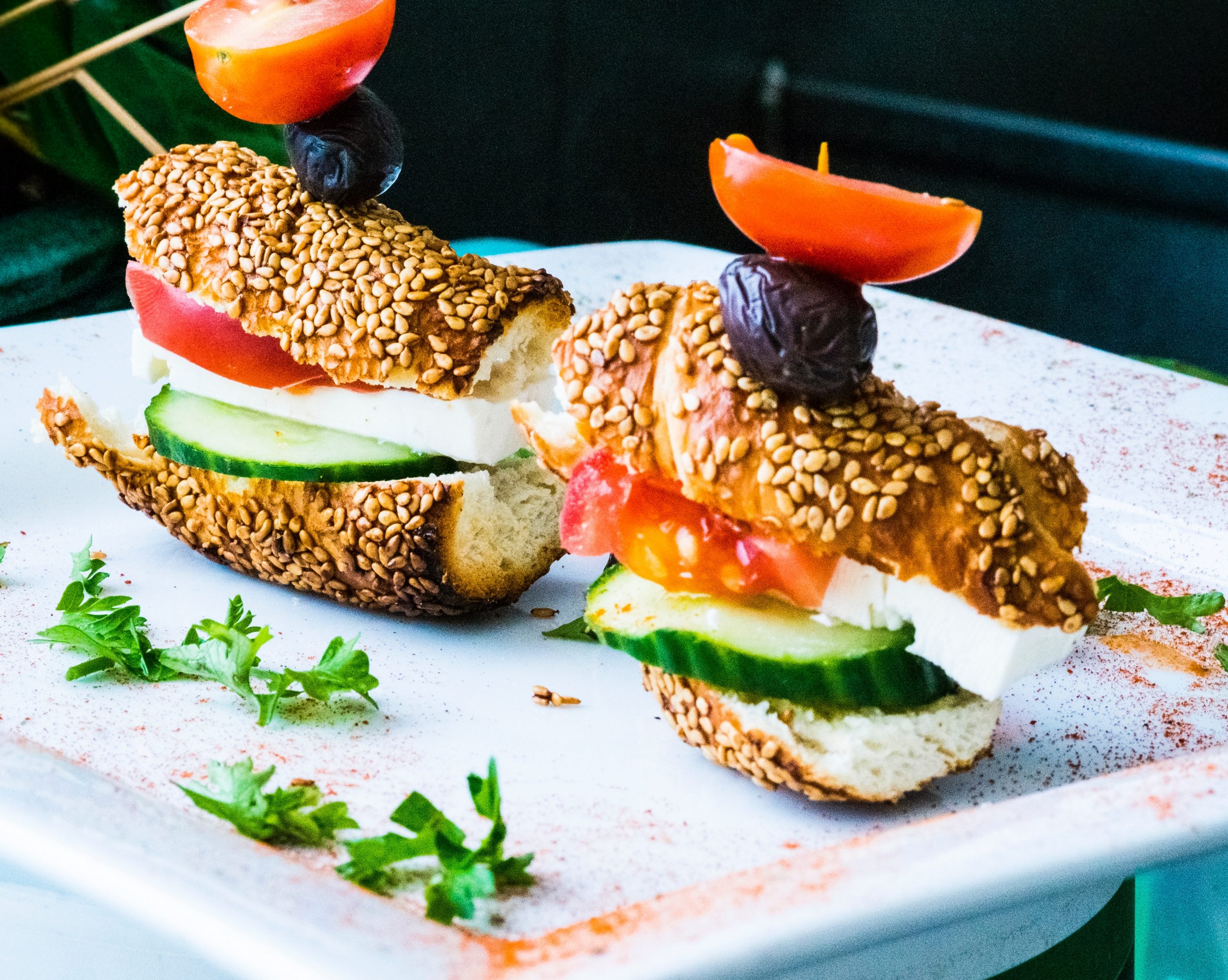 Zwei Sesamröllchen mit Feta, Gurke und Tomate, belegt mit Oliven auf einem weißen Teller, garniert mit Kräutern. Perfekt als köstliche Geschenkidee für jemanden, der den mediterranen Touch liebt!