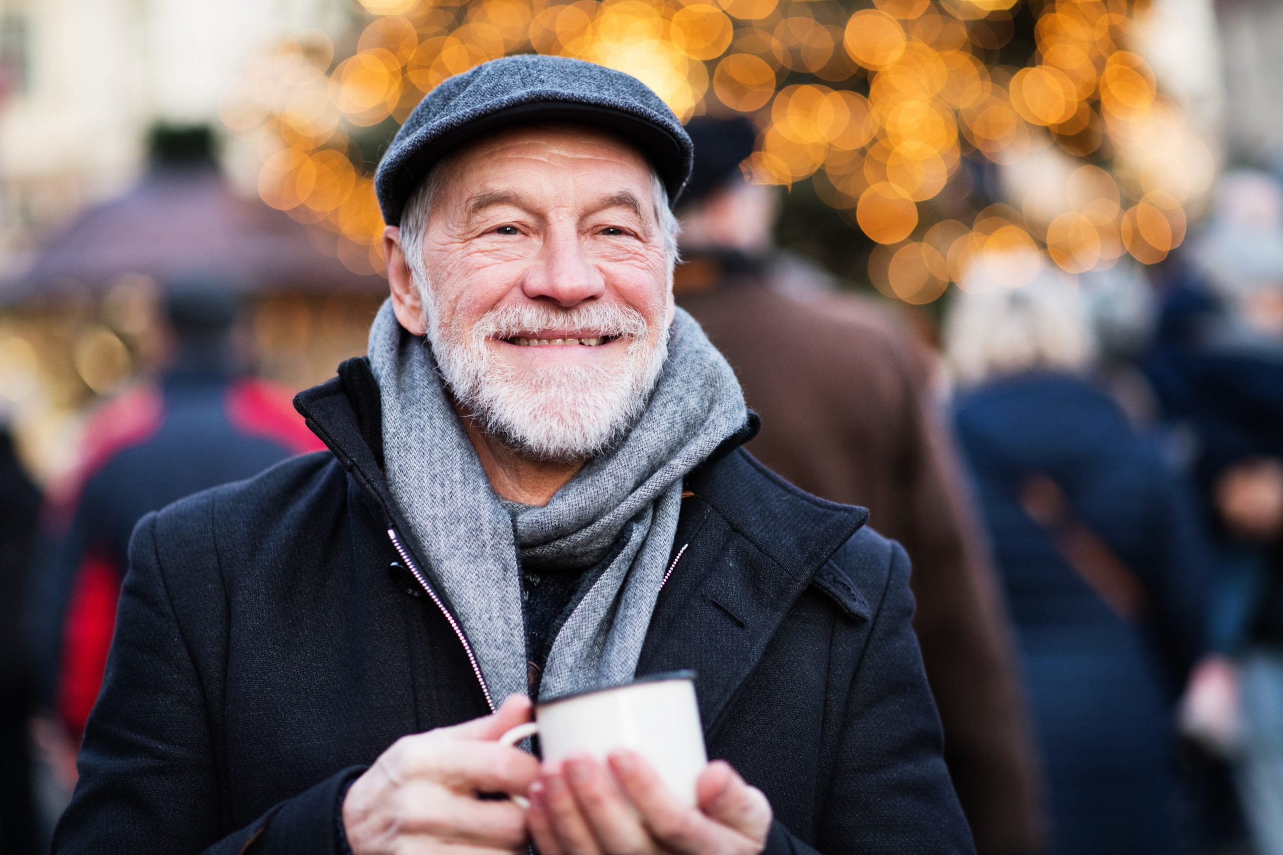 Ein älterer Mann in Mantel und Mütze lächelt herzlich und hält eine dampfende Tasse im Freien, umgeben von festlichen Lichtern. Seine Anwesenheit verkörpert den Geist der Freude und des Schenkens – eine perfekte Geschenkidee für diese Weihnachtszeit.