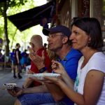 Menschen genießen ihr Essen im Freien auf einer Stadtstraße, sitzen auf einer Bank mit Bäumen und Fußgängern im Hintergrund – eine perfekte Kulisse für alle, die nach einer einzigartigen Geschenkidee suchen.