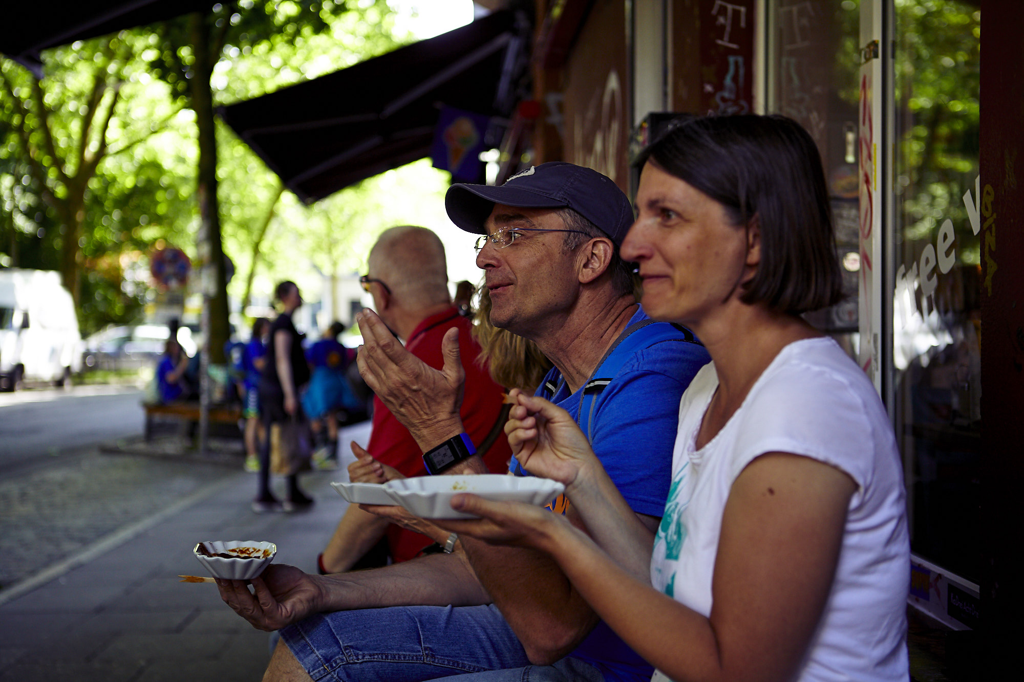 Menschen genießen ihr Essen im Freien auf einer Stadtstraße, sitzen auf einer Bank mit Bäumen und Fußgängern im Hintergrund – eine perfekte Kulisse für alle, die nach einer einzigartigen Geschenkidee suchen.