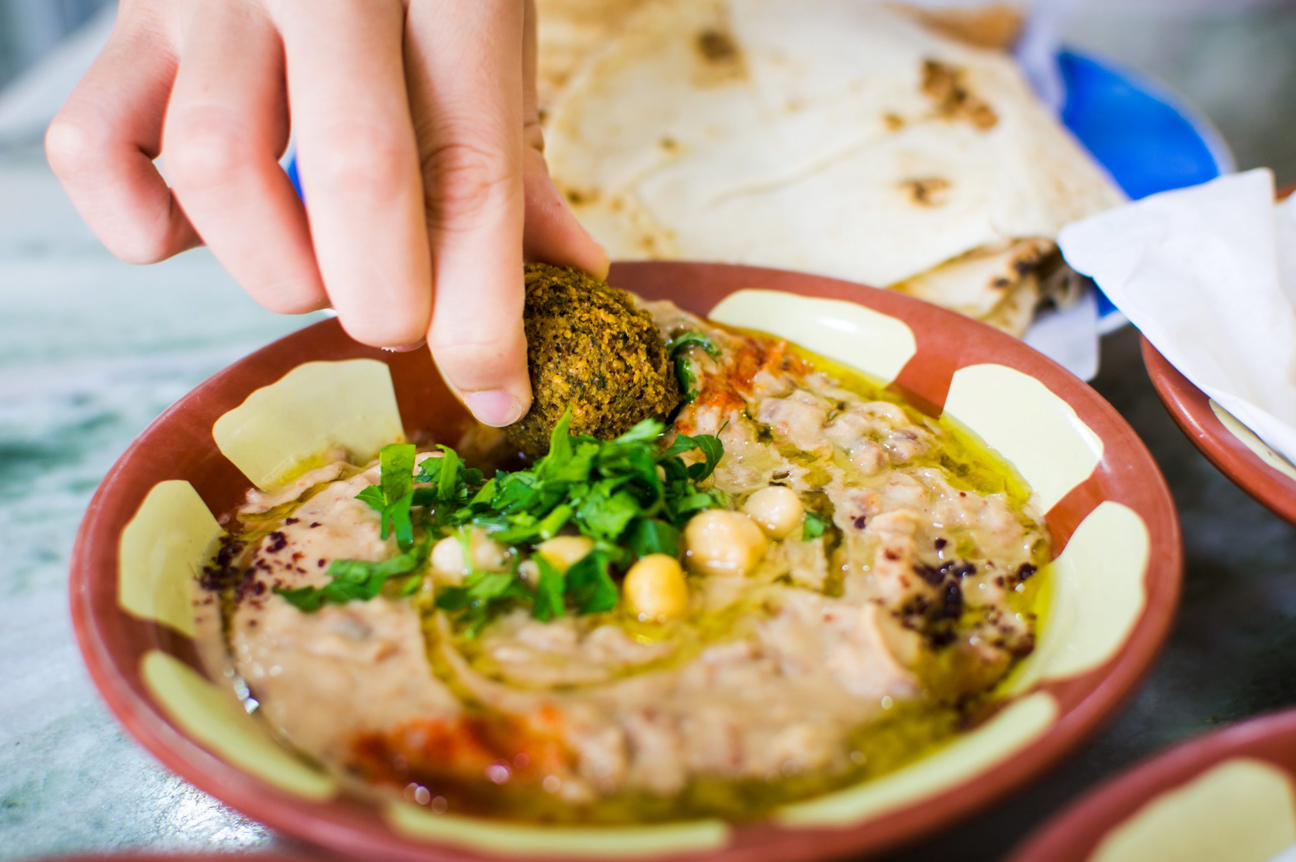 Falafel werden mit der Hand in eine Schüssel Hummus getaucht, garniert mit Petersilie, Kichererbsen und Olivenöl, im Hintergrund Brot – wirklich eine köstliche Geschenkidee für jeden Feinschmecker.