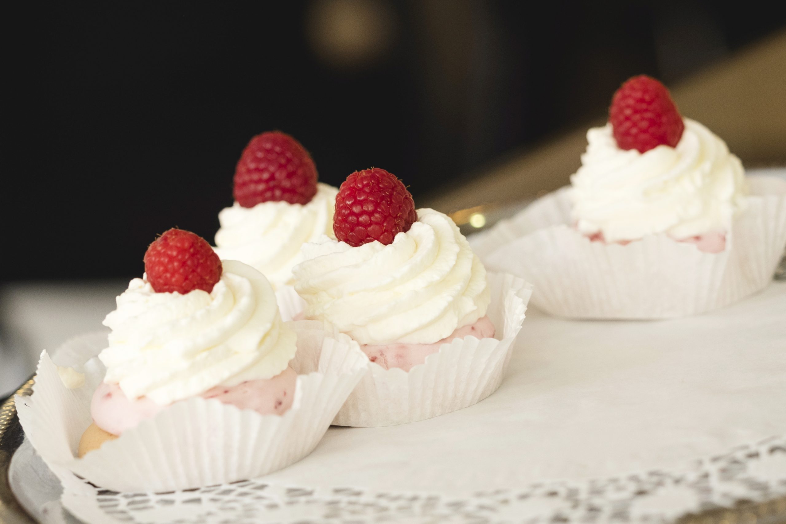 Vier mit Schlagsahne und Himbeeren garnierte Cupcakes ruhen elegant auf einem Tablett mit Spitzenpapier und sind eine entzückende Geschenkidee.