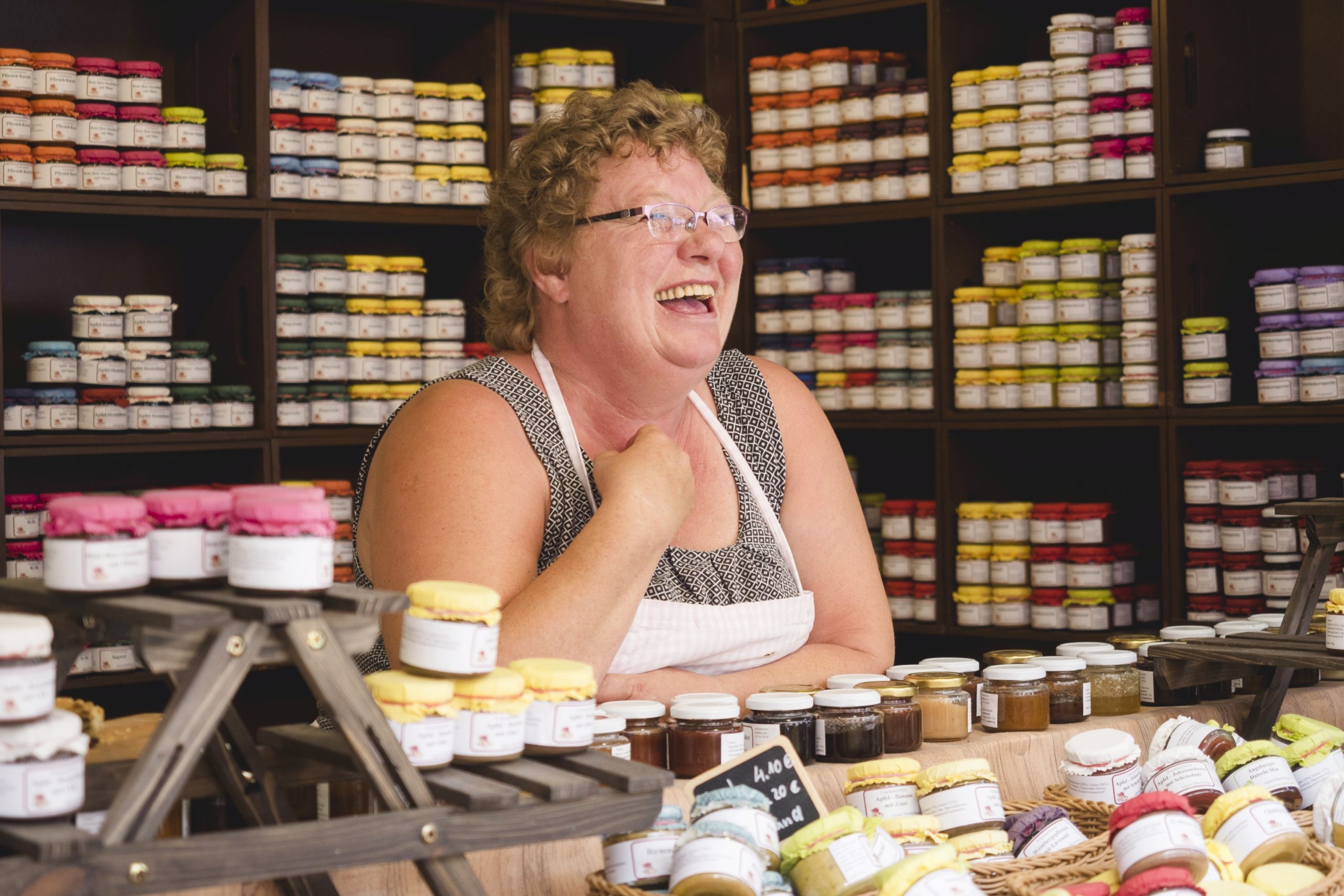 Eine lächelnde Person in einer Schürze steht hinter einer Theke voller bunter Gläser und Süßigkeiten, wahrscheinlich eine bezaubernde Geschenkidee. Mit Gläsern vollgestopfte Regale bilden einen lebendigen Hintergrund und tragen zur skurrilen Atmosphäre bei.