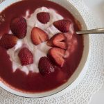 Eine Schüssel Joghurt, garniert mit geschnittenen Erdbeeren und einem Schuss roter Soße, steht elegant auf einer Unterlage mit weißem Spitzenmuster – wirklich eine entzückende Geschenkidee für jeden, der die Schönheit in der Einfachheit zu schätzen weiß.