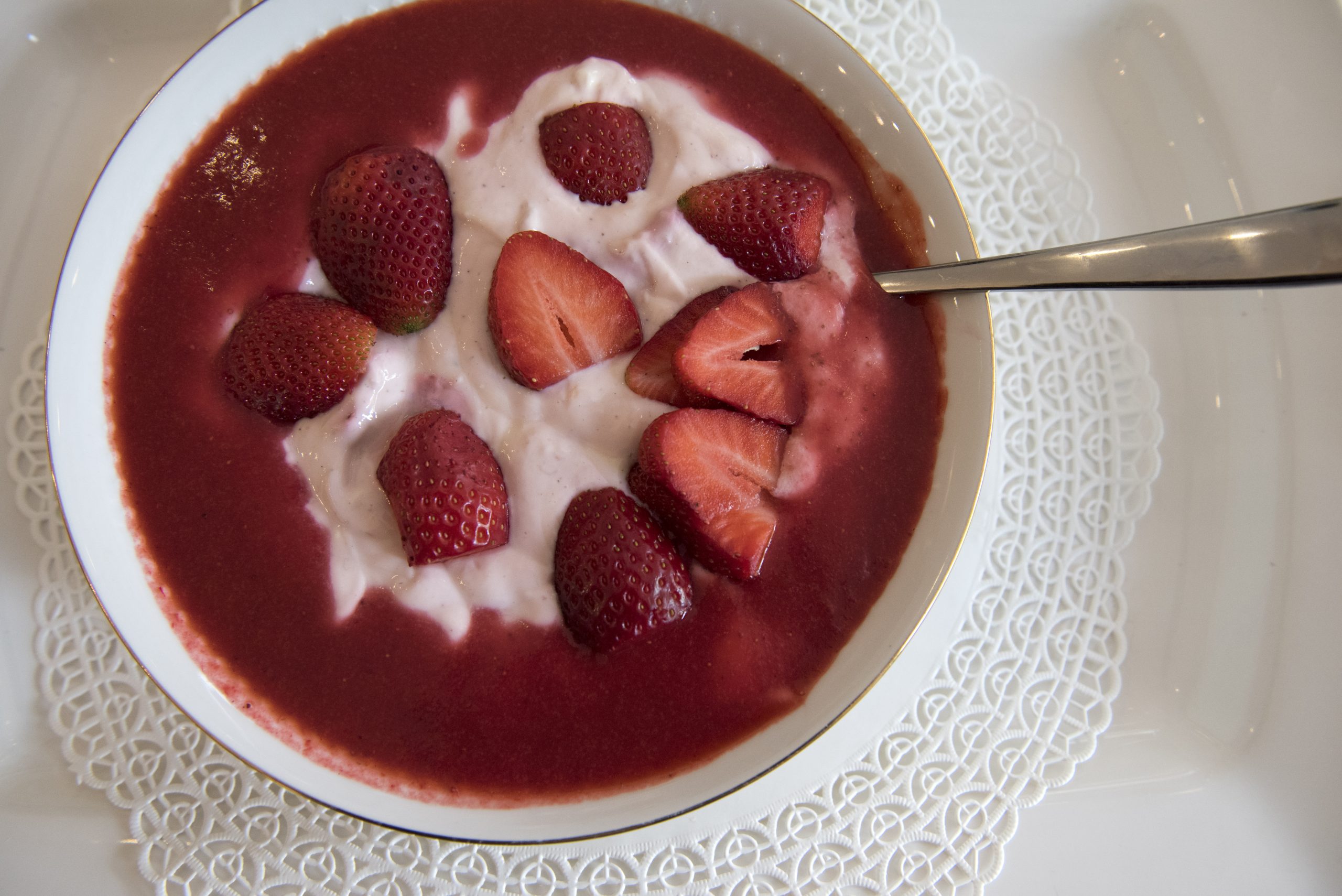 Eine Schüssel Joghurt, garniert mit geschnittenen Erdbeeren und einem Schuss roter Soße, steht elegant auf einer Unterlage mit weißem Spitzenmuster – wirklich eine entzückende Geschenkidee für jeden, der die Schönheit in der Einfachheit zu schätzen weiß.