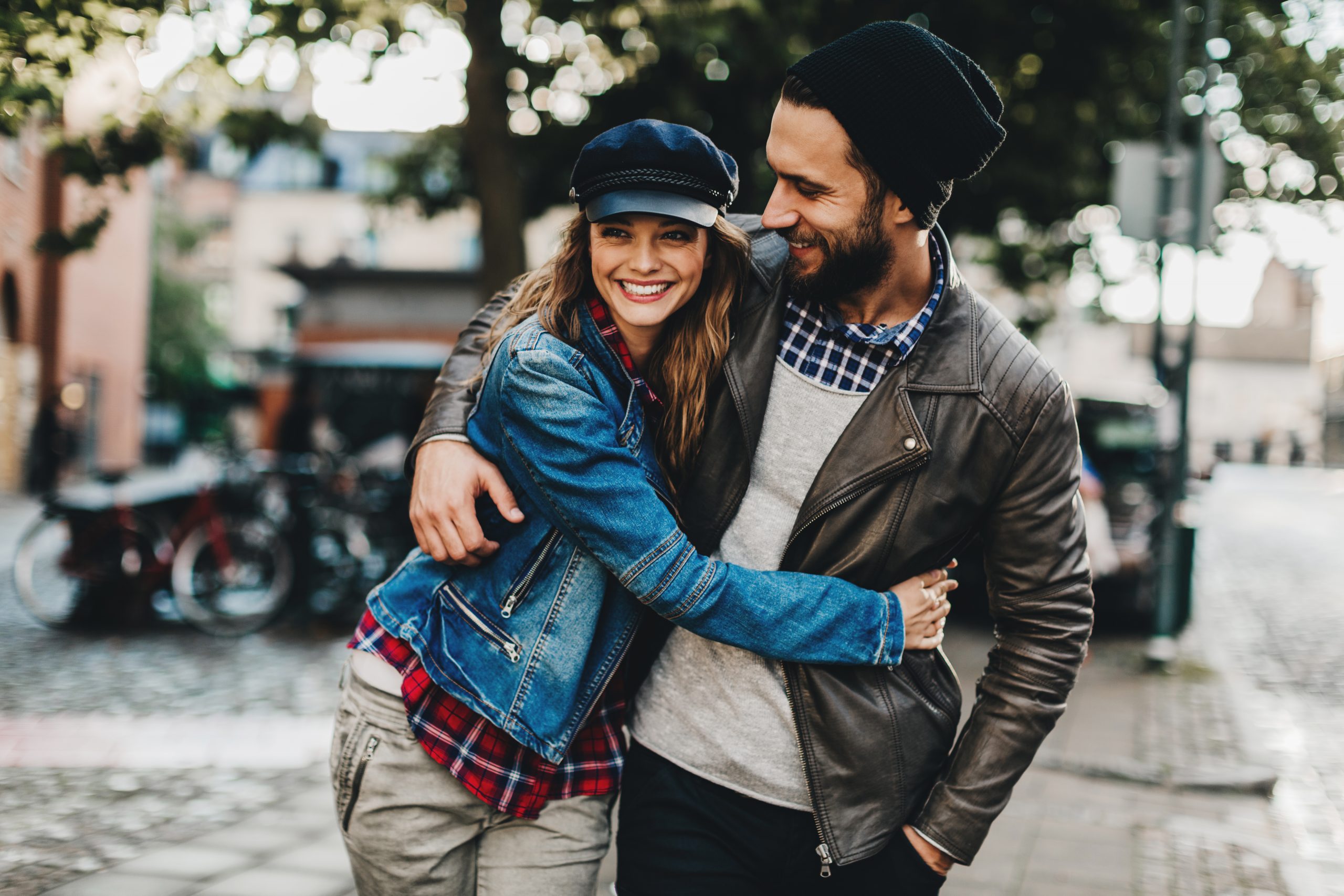 Ein glückliches Paar schlendert Arm in Arm über eine Kopfsteinpflasterstraße. Beide tragen legere Jacken und Hüte und lächeln, als ob ihr Tag voller Liebe wäre – und vielleicht die perfekte Geschenkidee.