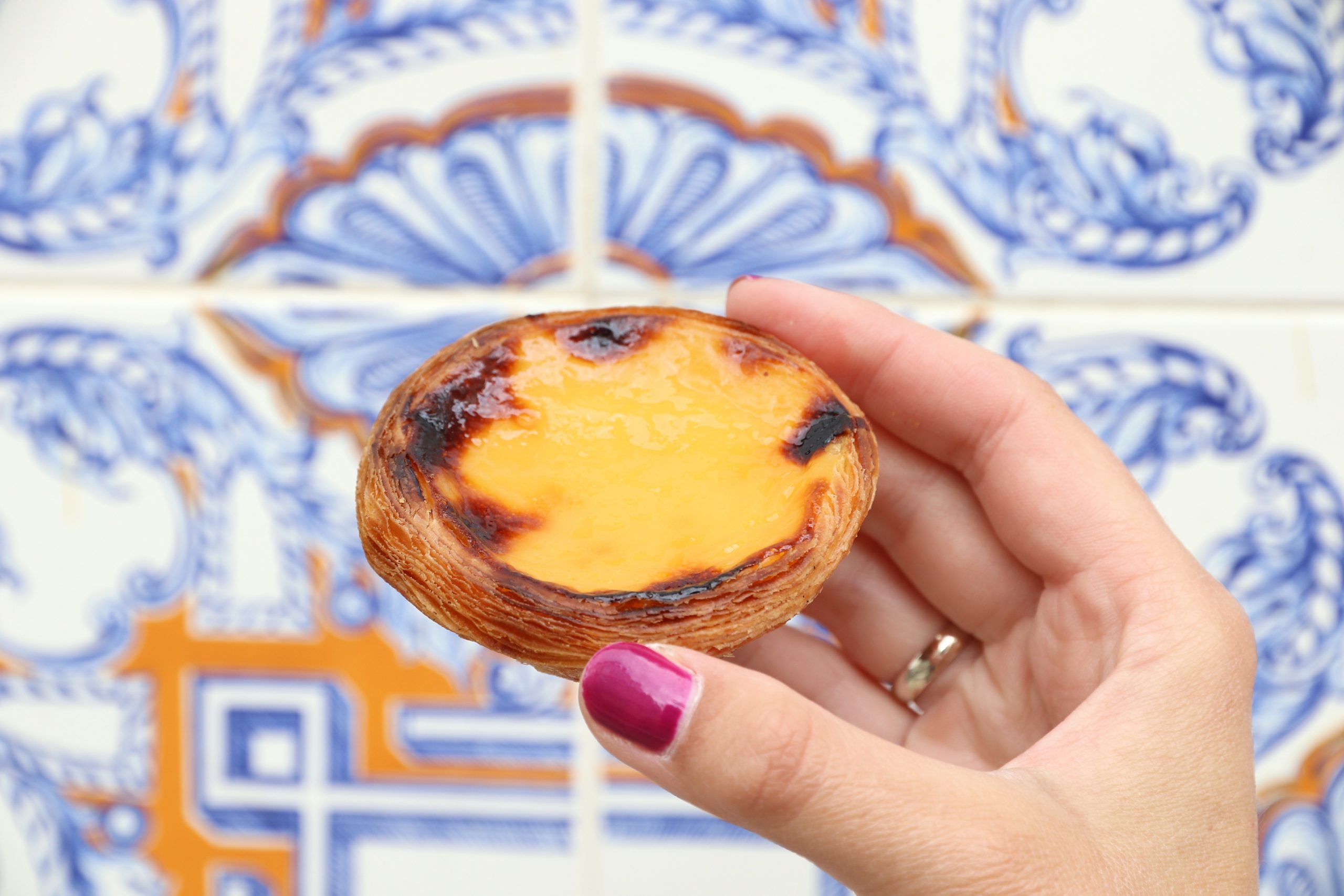 Eine Hand, die ein Pastel de Nata vor blauen und orangefarbenen Dekorfliesen hält, versprüht einen exotischen Charme und ist damit eine wunderbare Geschenkidee für alle, die kulinarische Kunst zu schätzen wissen.