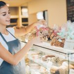 Eine lächelnde Frau in einer Schürze arrangiert verpackte Backwaren, jedes davon eine perfekte Geschenkidee, in einer Bäckerei-Vitrine.