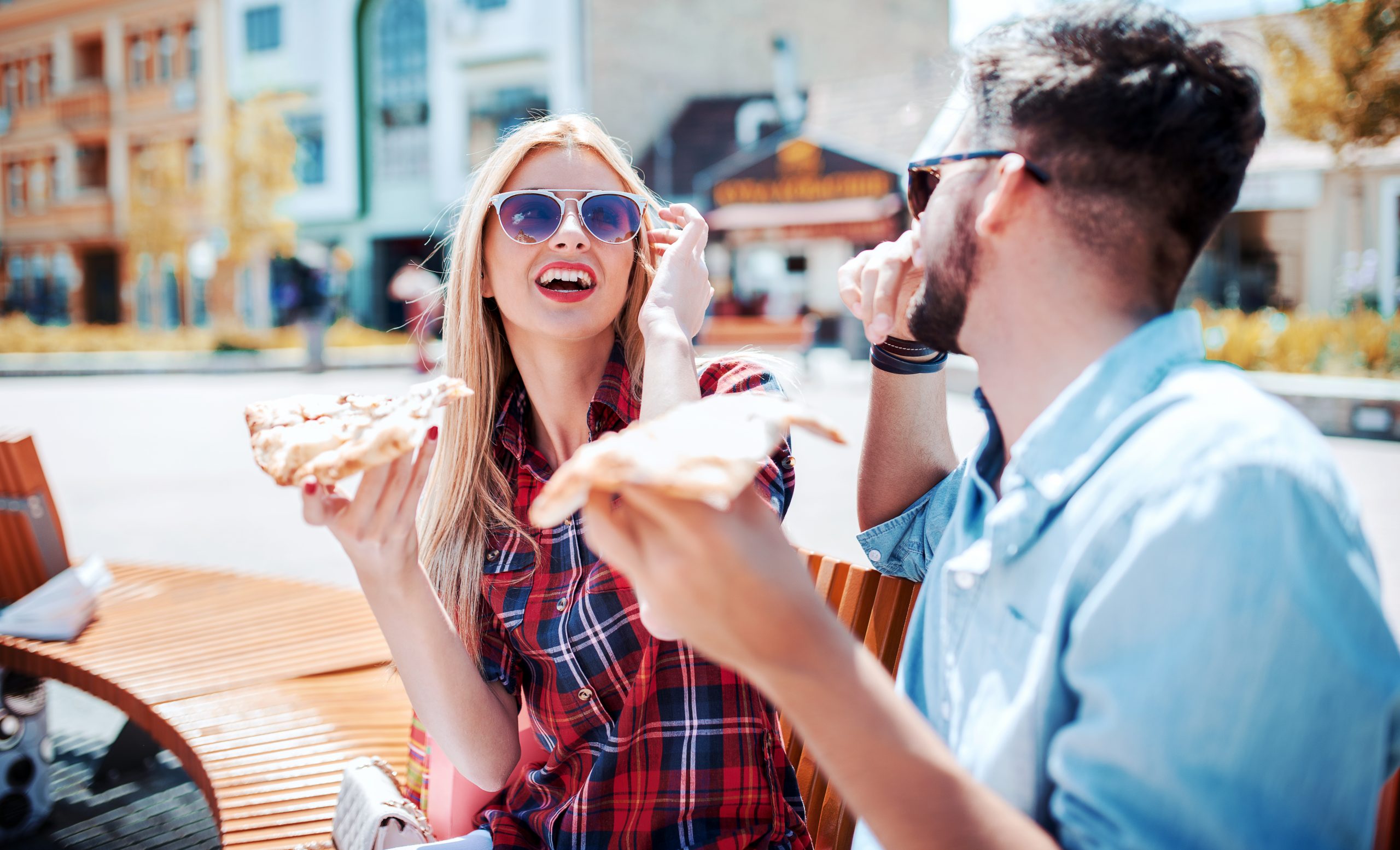 Zwei Menschen sitzen glücklich auf einer Bank im Freien, essen Pizza und tragen Sonnenbrillen – eine perfekte Geschenkidee für einen sonnigen Tag.