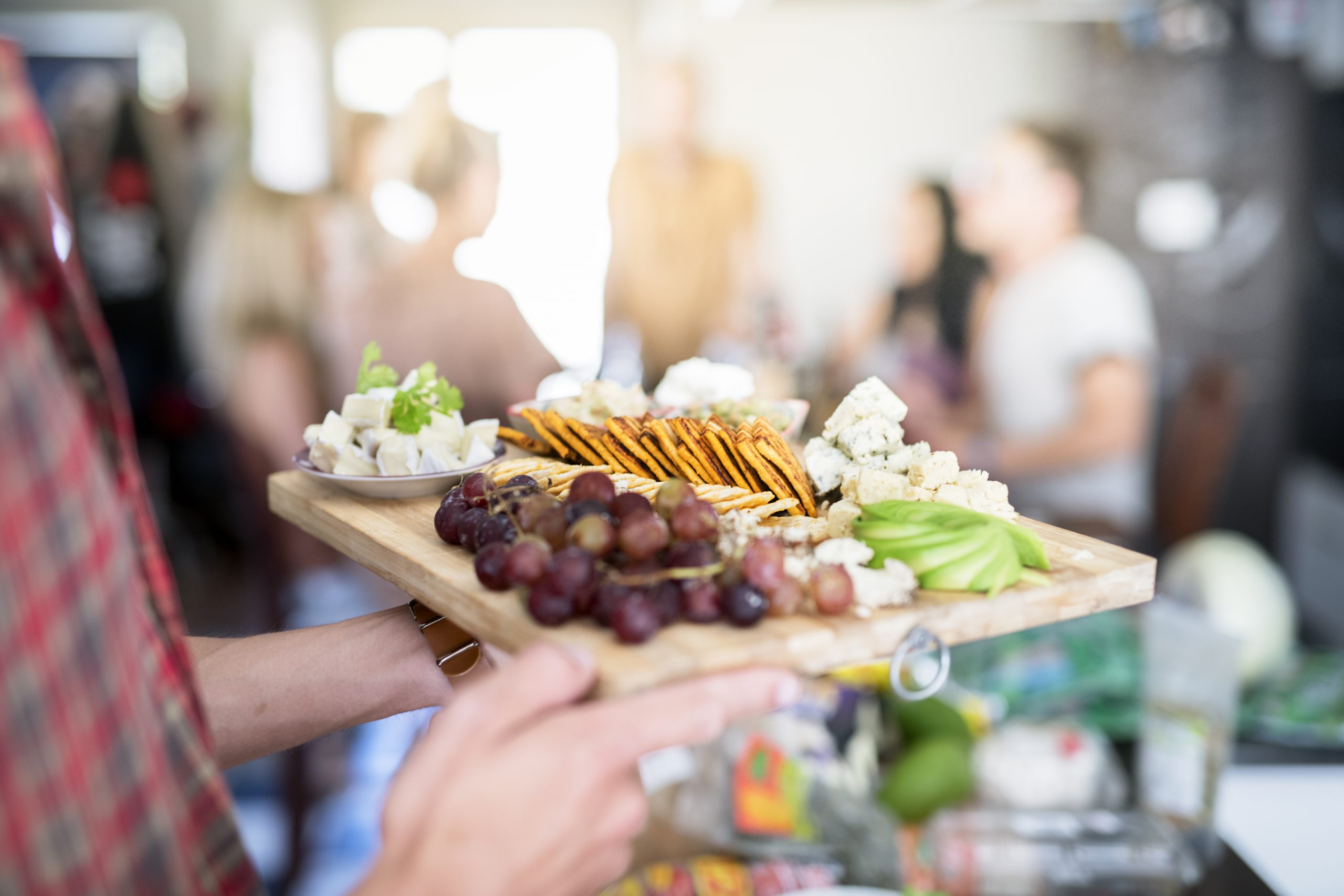 Bei einer Party hält eine Person eine Wurstplatte voller Trauben, Brezeln, Käse und Gemüse in der Hand – eine perfekte Geschenkidee für jeden Anlass.