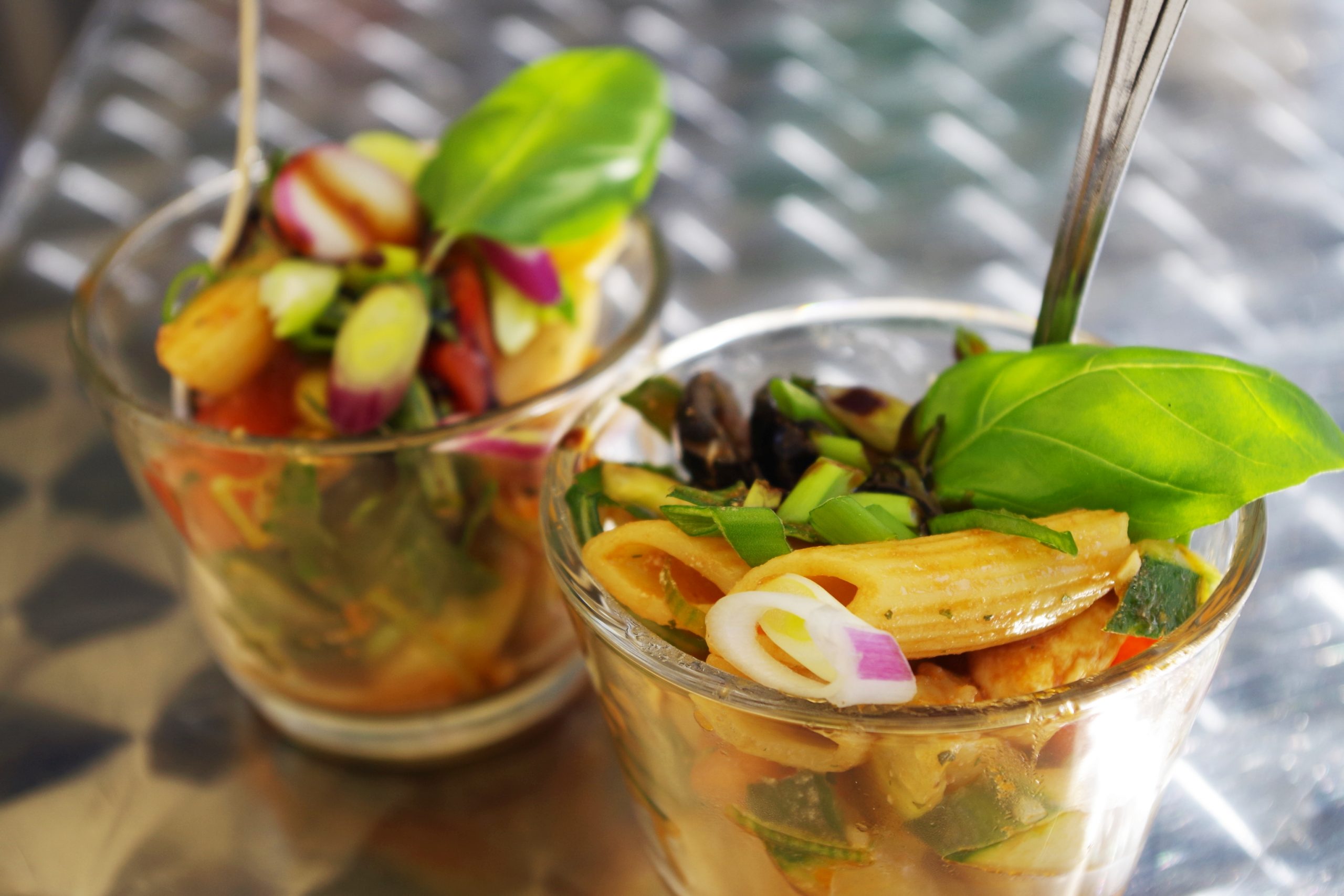 Zwei Gläser gefüllt mit Nudelsalat, garniert mit Basilikum, Frühlingszwiebeln und Radieschenscheiben, ruhen elegant auf einer reflektierenden Oberfläche – eine perfekte Geschenkidee für kulinarische Enthusiasten.