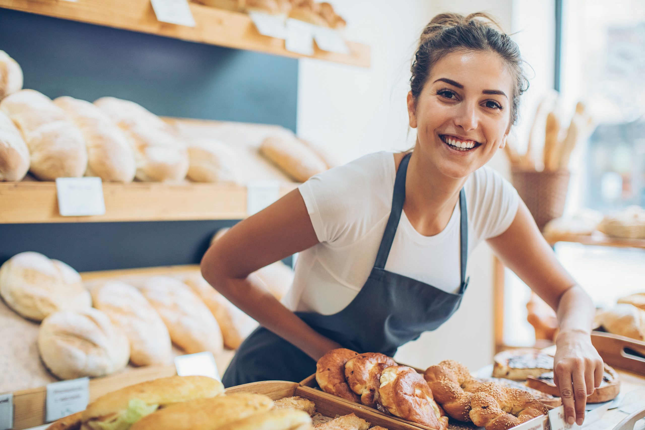 Eine lächelnde Frau in einer Schürze steht an einer Bäckereitheke, wo verschiedene Brot- und Gebäcksorten ausgestellt sind – perfekt als schöne Geschenkidee für jeden Anlass.