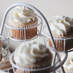 Cupcakes mit weißem Zuckerguss, elegant präsentiert auf einem weißen Metallständer vor einem hellen Hintergrund – perfekt als wunderbare Geschenkidee für jeden Anlass.