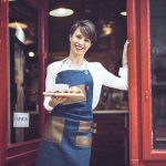 Eine Frau in einer Schürze hält mit einem herzlichen Lächeln ein Tablett mit Croissants an der Cafétür, die mit einem „Geöffnet“-Schild geschmückt ist – eine einladende Geste, die ihre Bäckerei zu einer wunderbaren Geschenkidee für jeden macht, der auf der Suche nach einem unvergesslichen Geschenk ist.