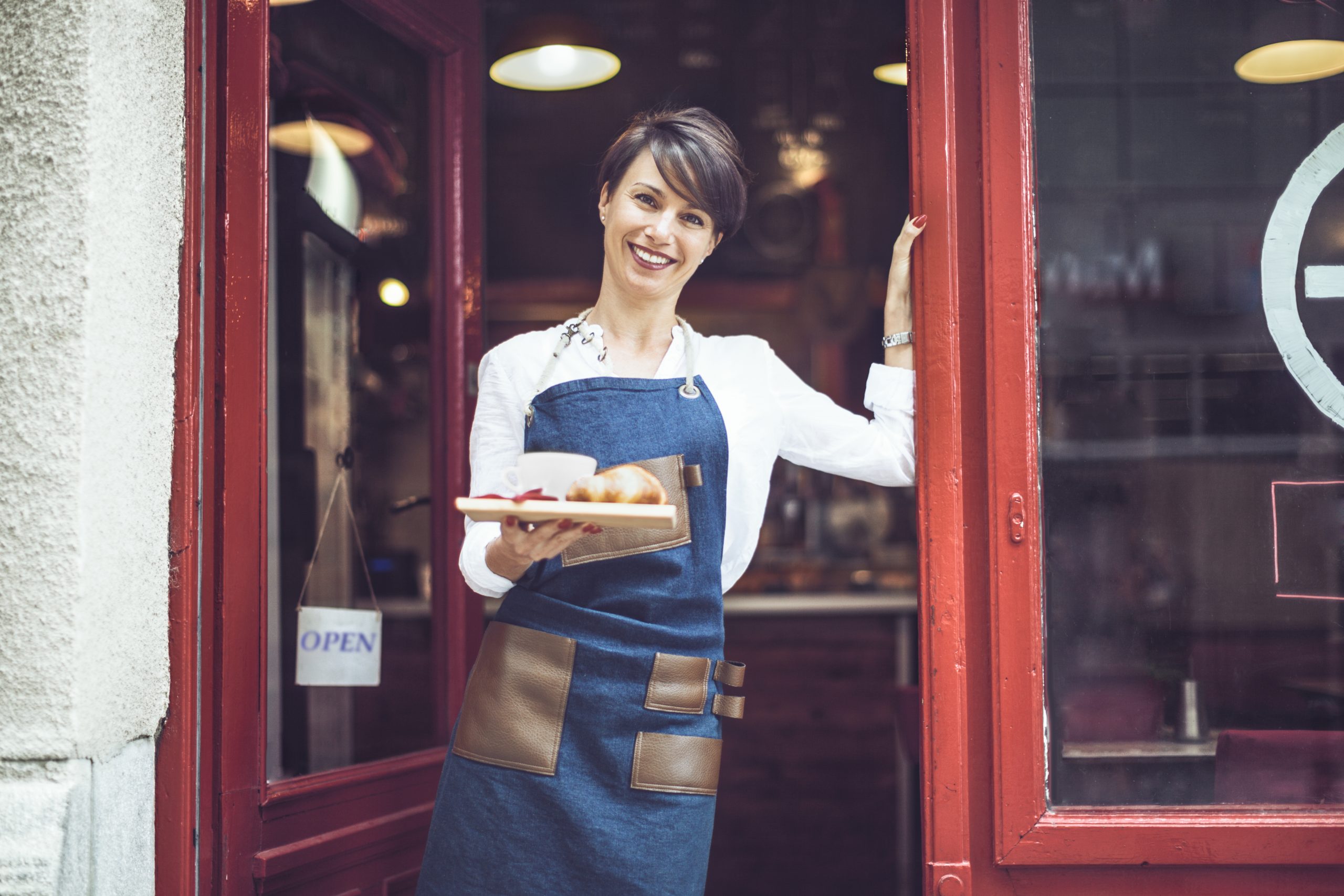 Eine Frau in einer Schürze hält mit einem herzlichen Lächeln ein Tablett mit Croissants an der Cafétür, die mit einem „Geöffnet“-Schild geschmückt ist – eine einladende Geste, die ihre Bäckerei zu einer wunderbaren Geschenkidee für jeden macht, der auf der Suche nach einem unvergesslichen Geschenk ist.