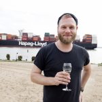 Mann hält ein Glas an einem Sandstrand, im Hintergrund ist ein großes Frachtschiff von Hapag-Lloyd zu sehen.