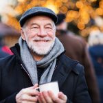 Ein lächelnder älterer Mann in Mantel und Mütze hält im Freien einen Becher, vor einem festlichen Hintergrund aus verschwommenen Lichtern und Menschen.