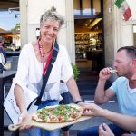 Eine lächelnde Person serviert Gästen im Freien in einem Restaurant Pizza, im Hintergrund sind eine Menütafel und eine italienische Flagge zu sehen.