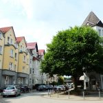 Straße mit farbenfrohen europäischen Gebäuden und einem großen Baum, geparkten Autos und einem Fahrradständer.