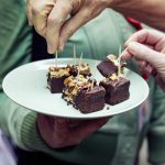 Hände greifen nach Schoko-Brownies mit Nüssen auf einem Teller, in jedem Stück ein Zahnstocher.