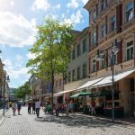 Eine belebte Kopfsteinpflasterstraße mit Menschen, Marktständen und historischen Gebäuden unter einem sonnigen Himmel.