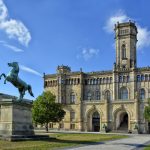 Statue eines sich aufbäumenden Pferdes vor einem historischen, reich verzierten Gebäude mit einem zentralen Turm an einem klaren Tag.