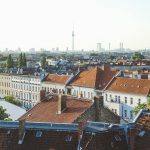 Stadtlandschaft mit Gebäuden mit roten Dächern und einem hohen Fernsehturm im Hintergrund unter klarem Himmel.