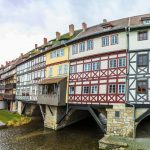 Eine Reihe farbenfroher Fachwerkhäuser auf einer Brücke über einen Fluss, im Hintergrund ein Kirchturm.